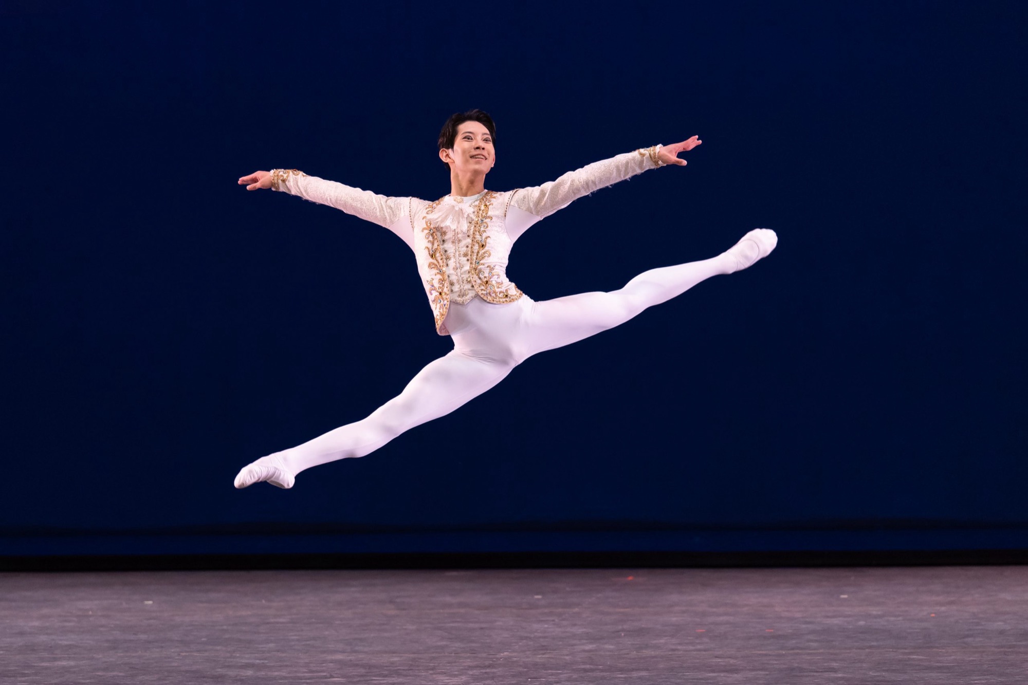 A male dancer in white and gold at the height of a split leap against a deep blue stage