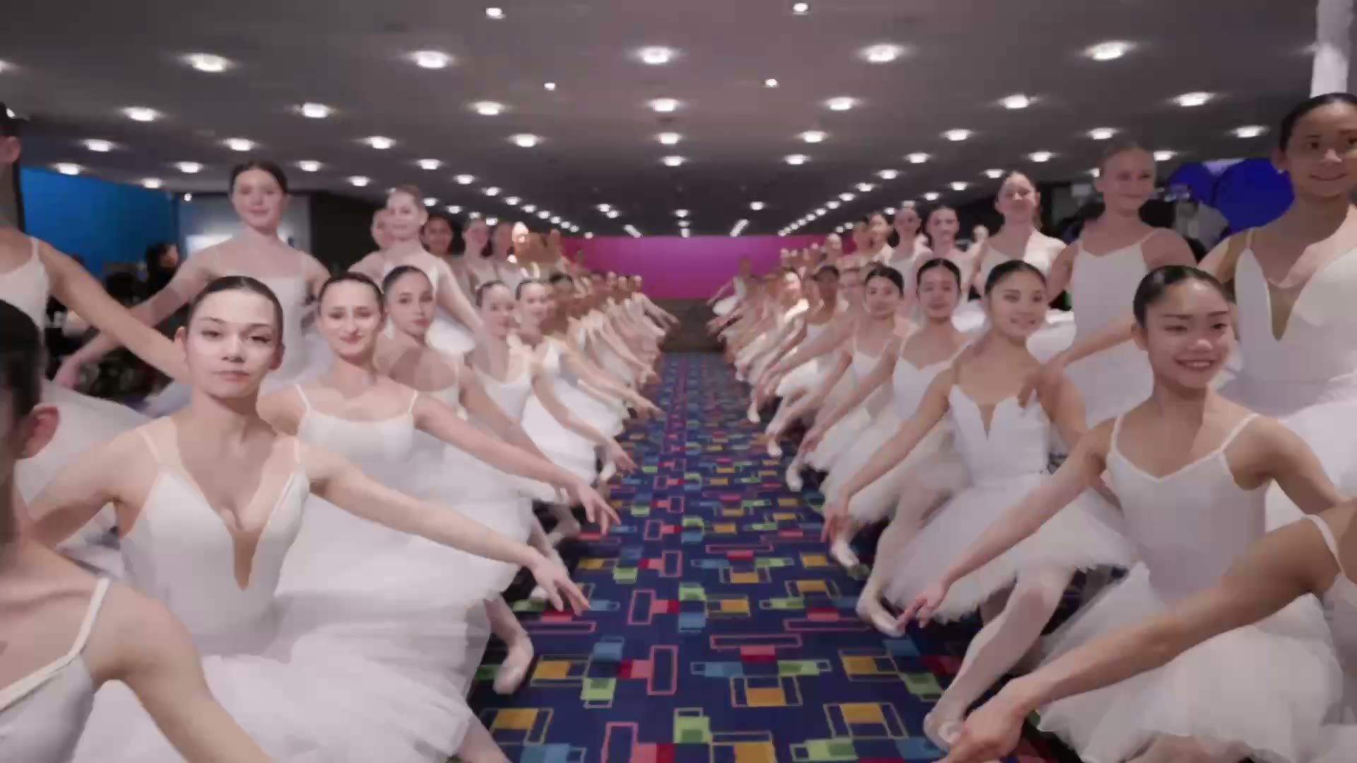 Rows of ballet students in white seated on the floor of a convention hall