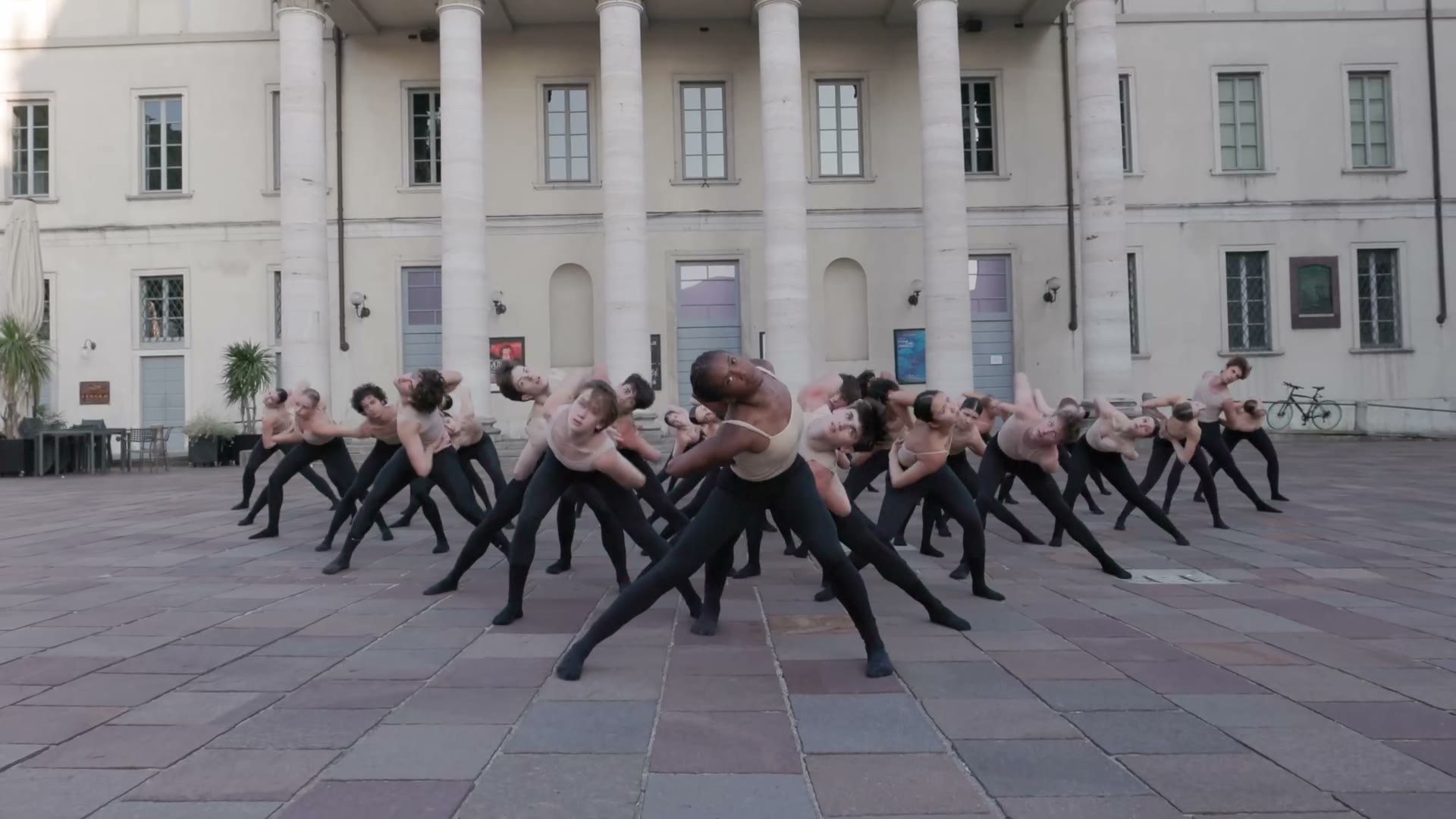 A company of dancers in lunges across a stone piazza in front of a columned building