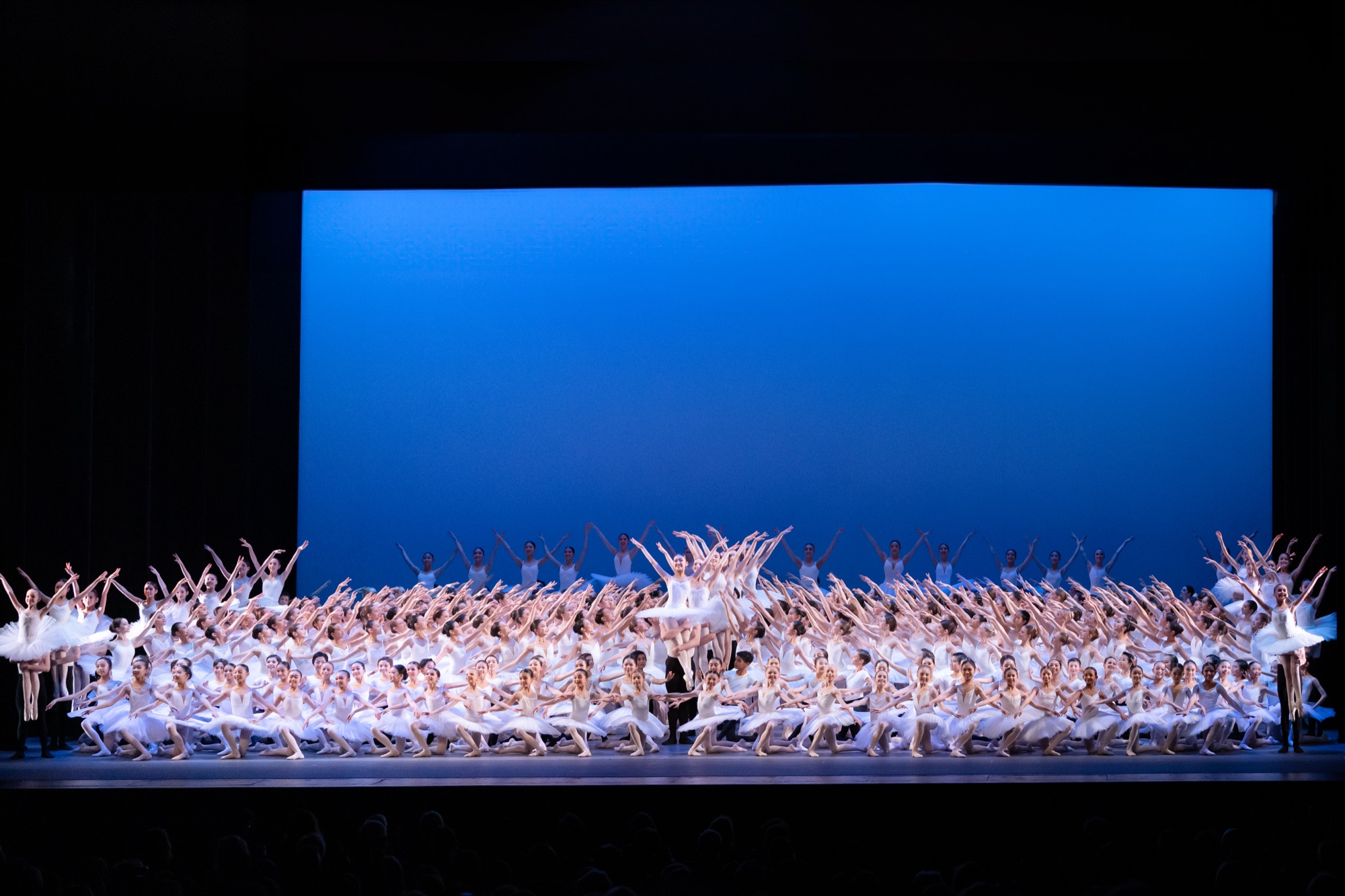 A full ballet company in white beneath a blue cyclorama