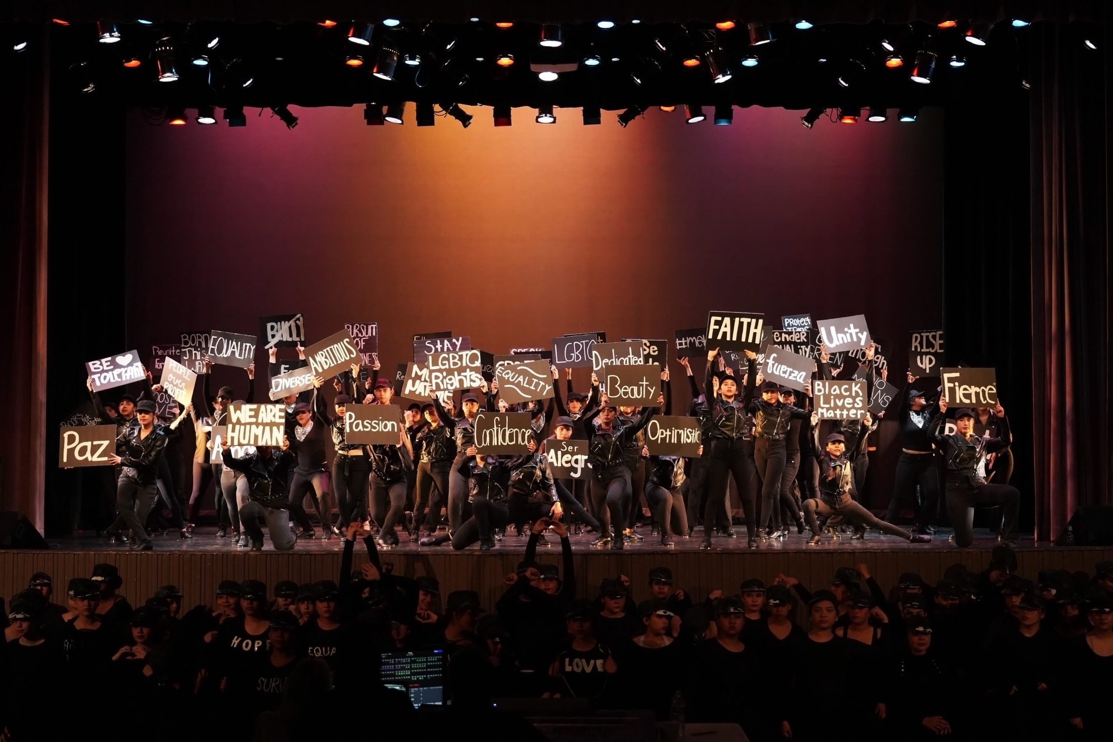 A full cast on stage at a performing arts event under warm light