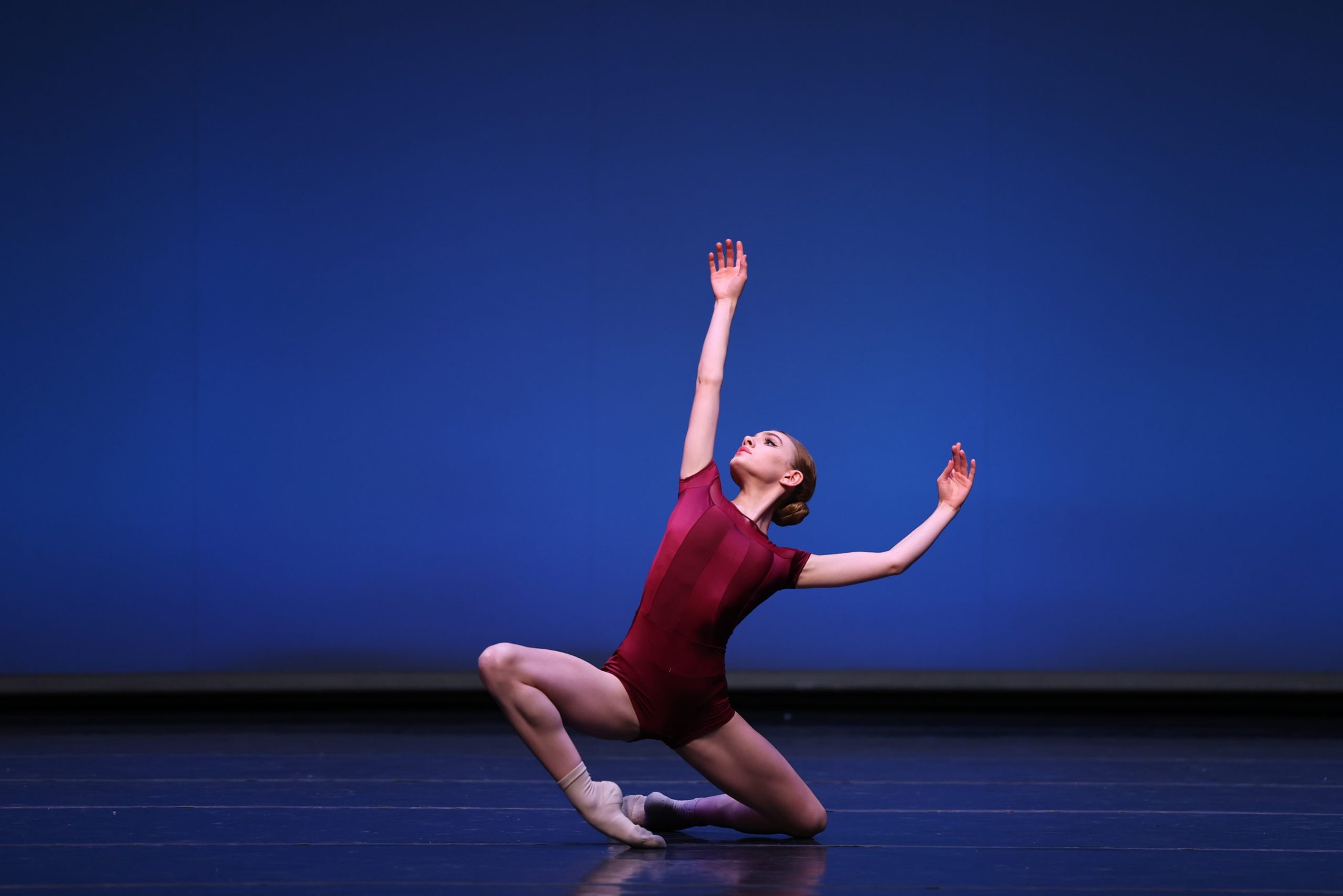 A contemporary dancer in burgundy kneeling with arms raised on a blue-lit stage